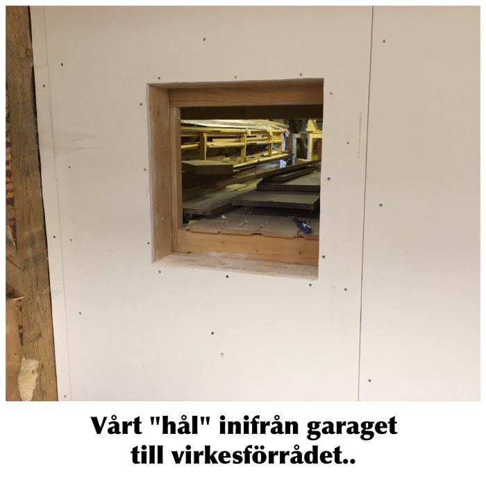 Hål i väggen inne i ett garage som leder till ett virkesförråd med längre brädor och reglar.