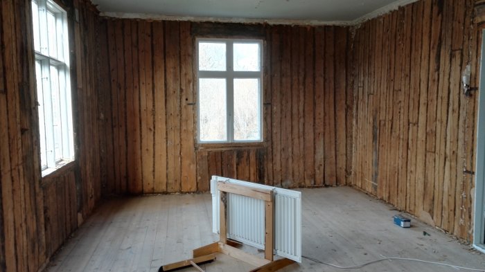 Interiör av ett renoveringsprojekt med synlig timmerstomme, fönster och en demonterad radiator.