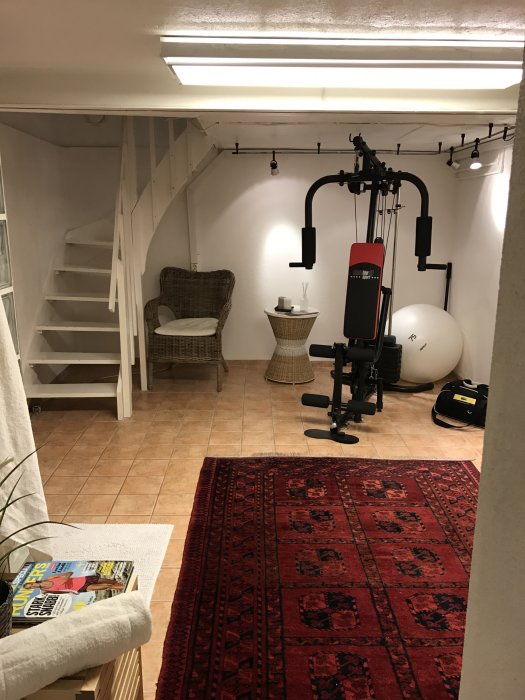 Källarrum med träningsutrustning, en korgstol och trappa, lågt tak med synliga rör och vitmålade väggar.