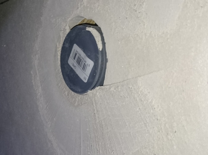 Avloppsanslutning täckt av plastlock monterad i vägg, med omgivande spackel och lite utstick.