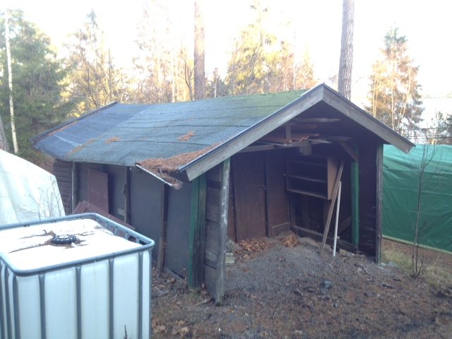 Nedgånget, övergivet garage med skadat tak och bristfälliga väggar omgivet av träd.