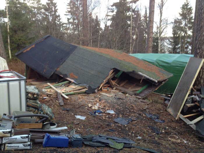 Ett fallfärdigt garage som kollapsat bland träd och skräp, med byggmaterial och verktyg utspridda på marken.