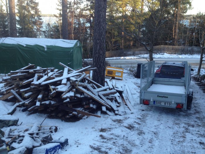 Hög med rivningsavfall från gammalt garage vid sidan av en släpvagn i snötäckt landskap.