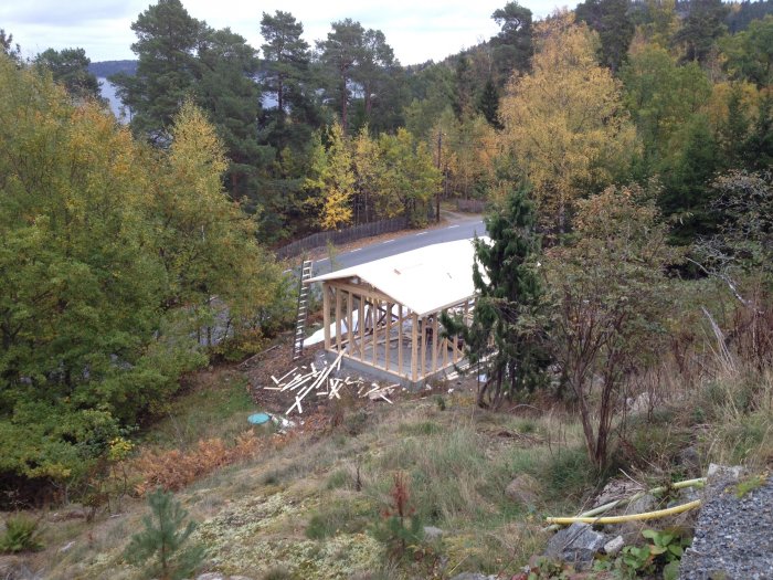 Konstruktion av garage med trästomme och tak på en grusig grund omgiven av skog och höstlöv.