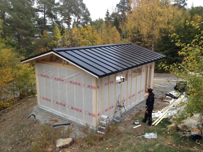 Nybyggt garage med takplåtar och vindskydd, omgivet av höstnatur och en person i förgrunden.