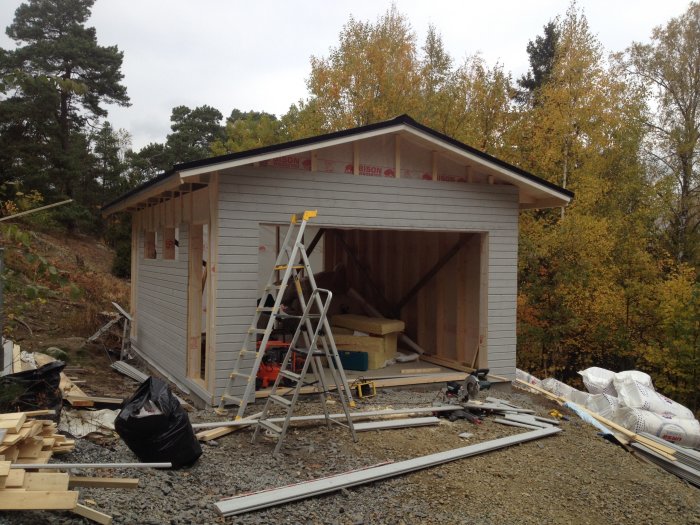 Nybyggt trägarage med grå panel och öppen port, byggmaterial och stege runtomkring, omgivet av höstträd.