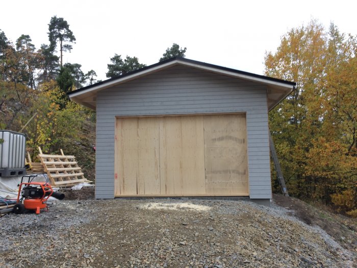 Nästan färdigbyggt gråmålat garage med plywoodtäckt port, omgivet av höstnatur och byggmaterial.