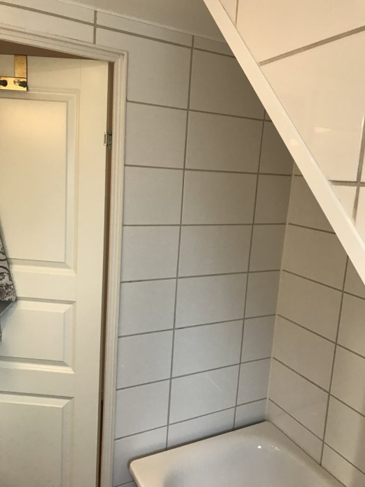 Badrum med vita kakelväggar och badkar under sluttande tak, dörr till vänster.