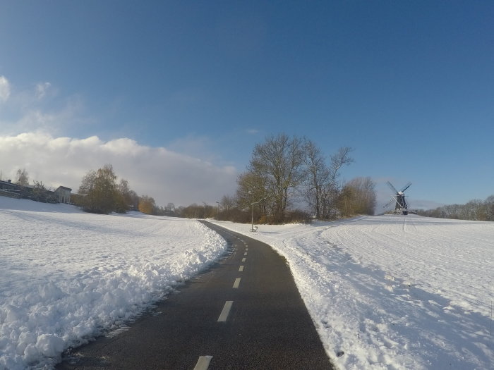 Vit vinterlandskap med snötäckt väg och en väderkvarn i bakgrunden under en klarblå himmel.