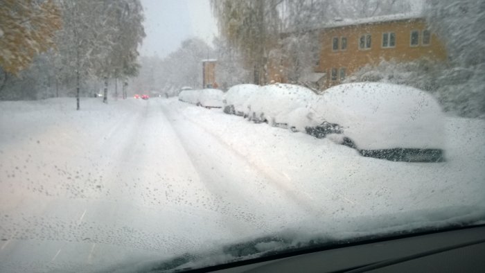 Vy från bil på snöig dubbelriktad väg i Bromma med parkerade bilar täckta av snö.