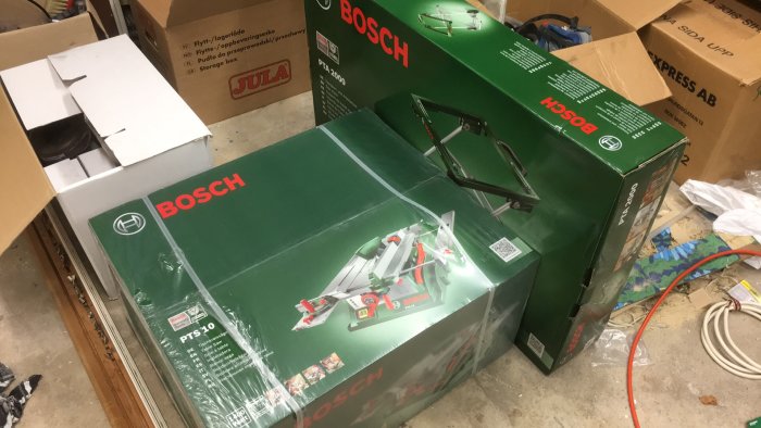 Nya Bosch PTS-10 bordcirkelsåg och PTA 2000 rullbord i förpackningar på ett stökigt garagegolv.