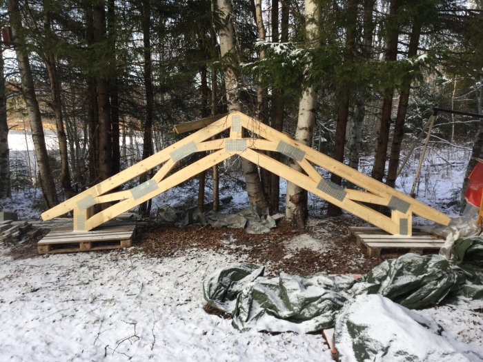 Nya takstolar ligger på träpallar snötäckta på marken med skog i bakgrunden.