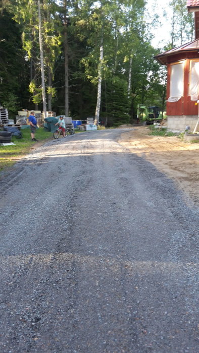 Grusväg på landsbygden med person och barn på cykel omgiven av träd och hus.