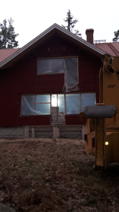 Röd lantlig villa under renovering med plasttäckta fönster och en grävmaskin i förgrunden.