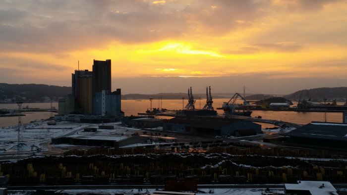 Solnedgång över grått industriområde i Uddevalla med orange himmel och silhuetter av byggnader och kranar.