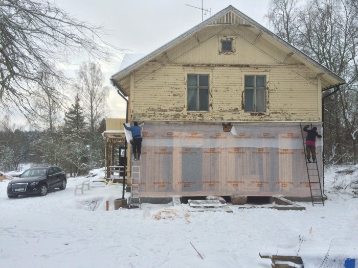 Renoveringsarbete på en gammal trävilla under vintern, vindpapp monterad på fasad, rivna betongtrappor och bil parkerad framför huset.