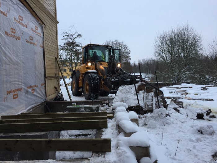 Hjullastare tar bort gamla betongtrappor vid en husgrund med synligt bjälklag och snötäckt mark.