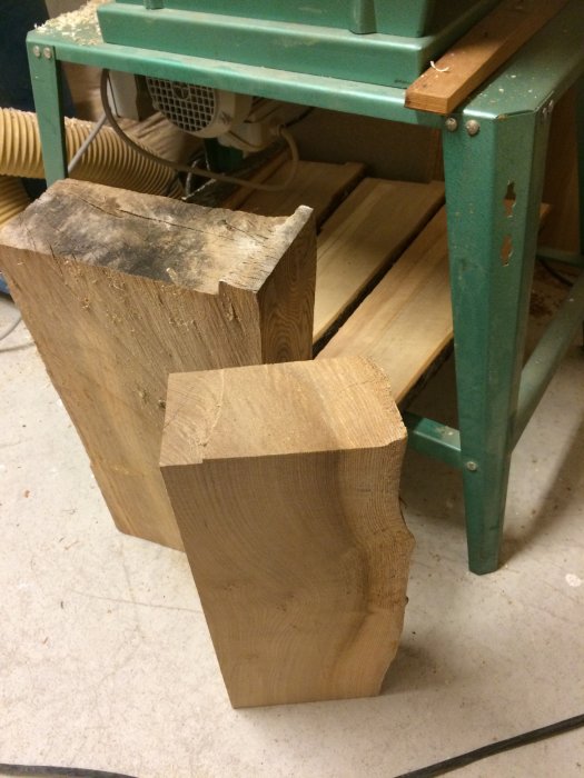 Två nysågade bitar av almträ på ett verkstadsgolv, nära en grönmålad arbetsbänk.