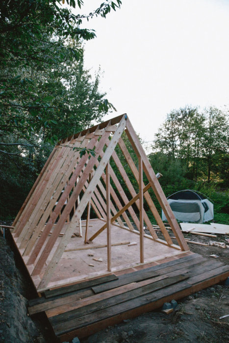 Trästommen till ett litet hus i byggnation med takreglar på en plattform av trä, omgivet av natur.