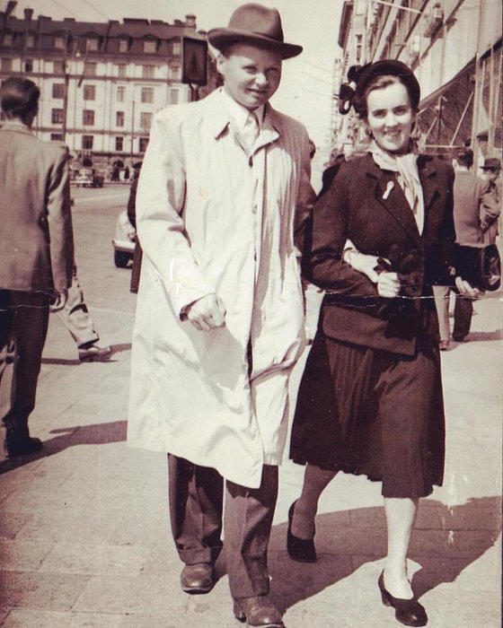 Ett äldre par klätt i typisk klädsel från 1950-talet promenerar på en gata i Stockholm.