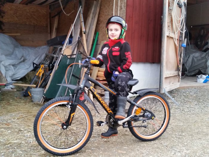 Barn i hjälm sitter på en orange och svart mountainbike framför ett öppet förråd fyllt med redskap och prylar.