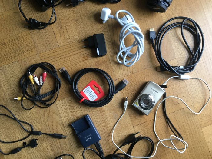 Olika elektroniktillbehör på ett golv: kablar, laddare och en digital kamera.