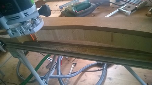 Verktyg fräser ett bågformat karmöverstycke i trä, fastsatt på arbetsbänk, med MDF-spån och verktyg synliga.