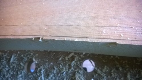 Nätbild på en träkarm delvis fräst med synliga flis och spån vid kanten, med hela skruvar i bakgrunden.