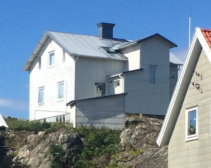 Ett vitmålat hus med frontespis och trapphus, beläget på en berghäll, omgivet av andra hus och grönska.