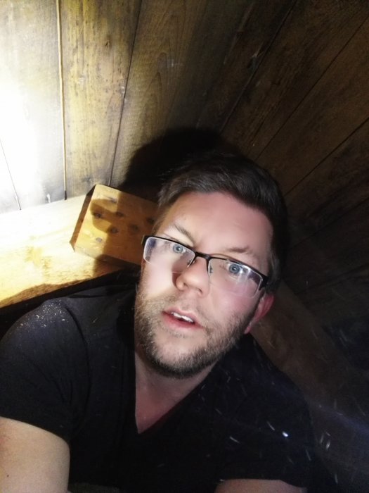 Man i trångt utrymme på vind med synliga träbjälkar och mörk bakgrund, ser ut att bedöma arbetet.