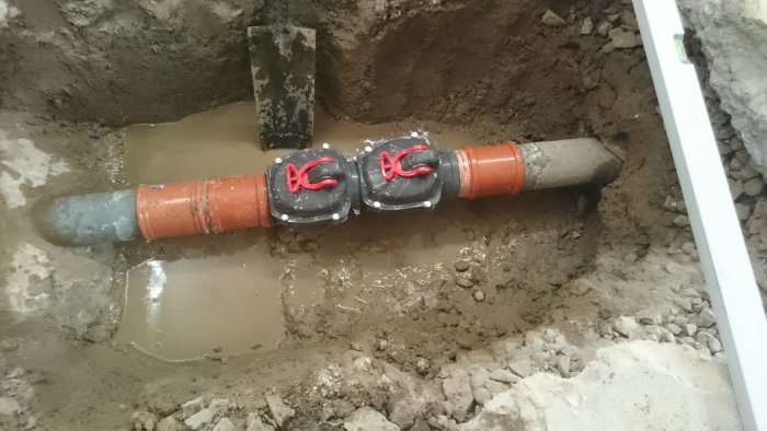 Grävd grop med två sammankopplade rörledningar och lera runtomkring under gjutningsarbete.