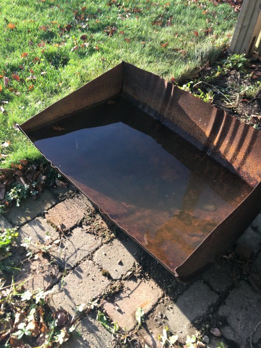 Rostig metallbehållare med vatten står på en stenlagd mark omgiven av gräs och löv.