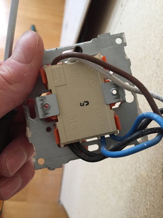 Hand som håller en demonterad strömbrytare med synliga kablar och klämmor vid en vägg.