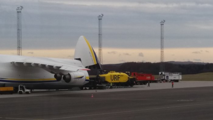 Gult objekt, möjligtvis en ubåt, lastas in i bakdelen på ett stort flygplan med siluetten av en dramatisk himmel i bakgrunden.