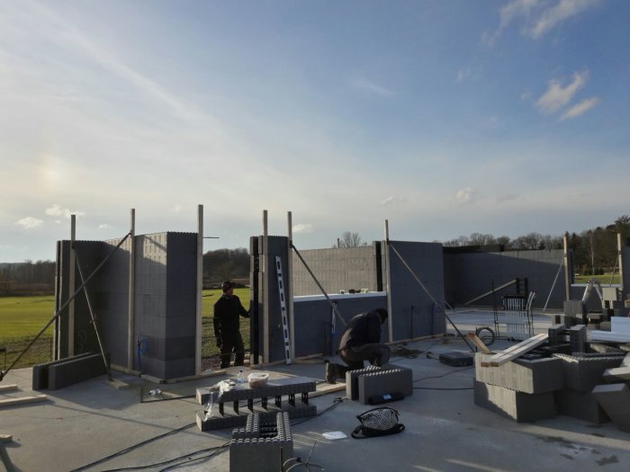 Två personer staplar betongblock för att bygga väggar på en husgrund under en blå himmel.