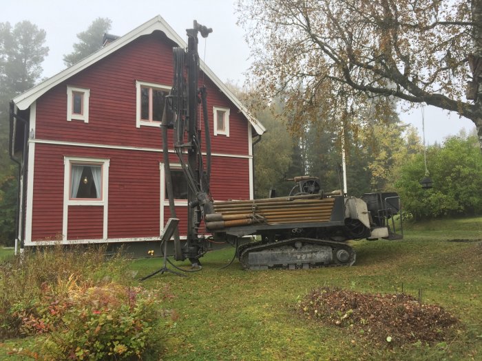 Borrigg i arbete framför rött hus med bergvärmeinstallation, kuperad trädgård i bakgrunden.