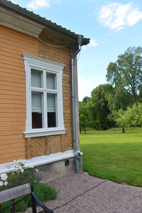 Hörn av gammalt hus med regnvattenrör och stänkskydd, före renovering, grönt gräs och träd i bakgrunden.