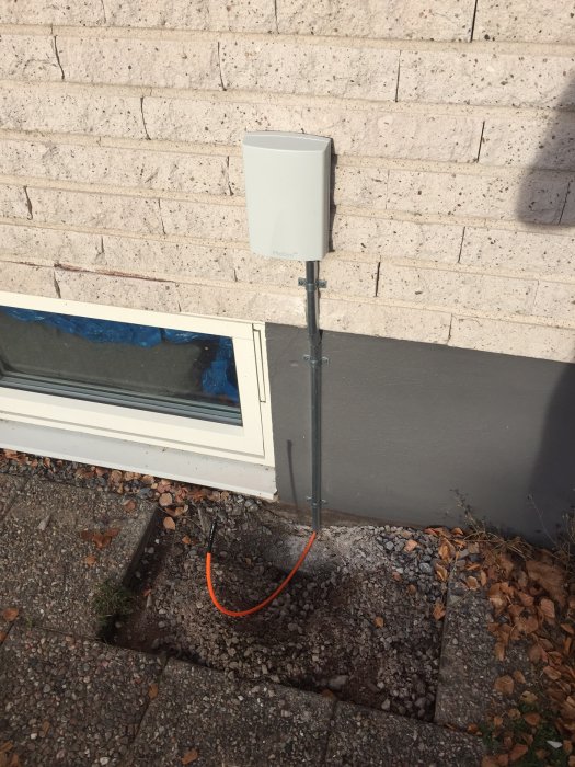 En kopplingslåda för fiberinstallation monterad på en husvägg med en synlig orangeröd kabel och en öppen grävd kanal i marken.