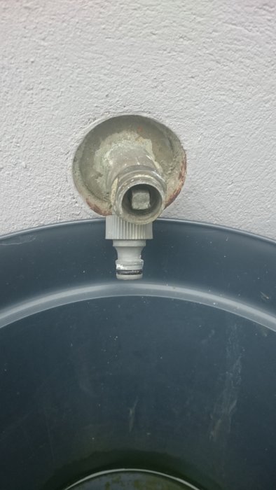 Rostig vattenutkastare installerad ovanför en svart plastbehållare mot en vit vägg.