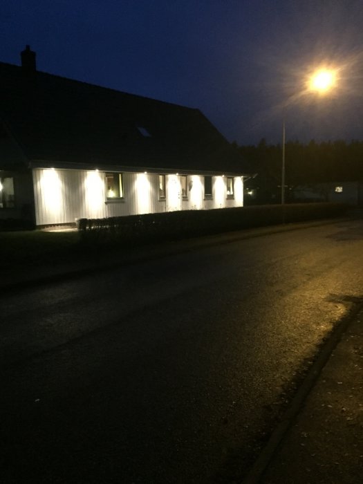 Ett upplyst enfamiljshus vid gryning med tända fönsterlampor och en tänd gatlykta.