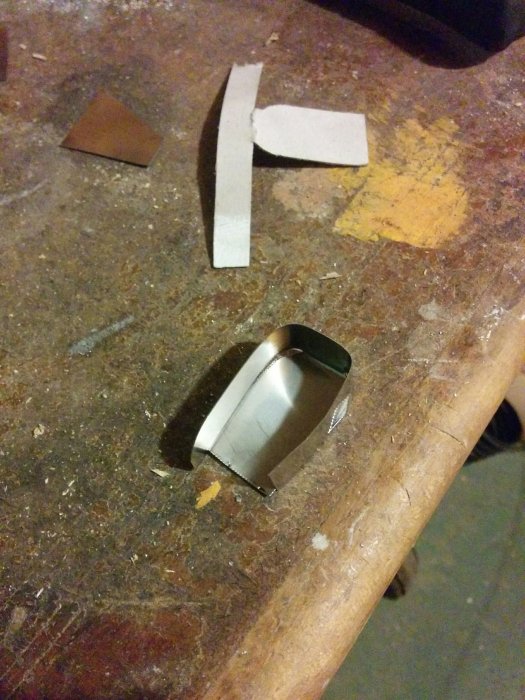 En metallbit med böjda kanter och en pappersmall ovanpå på en sliten verkstadsbänk, omgiven av verktyg och andra materialbitar.