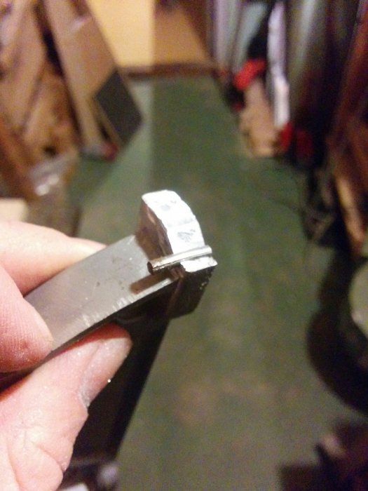 Hand håller en trasig aluminiumdel nära kameran i ett suddigt rum.