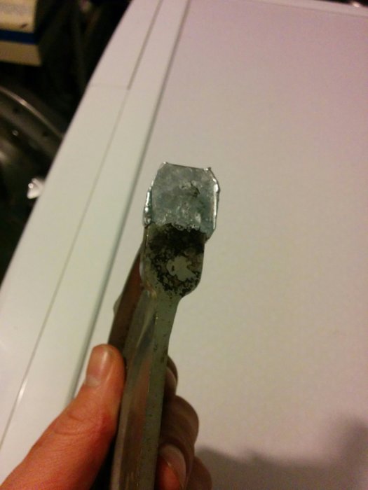 Hand som håller en metallstång med ett smält metallstycke i änden mot en vit bakgrund.