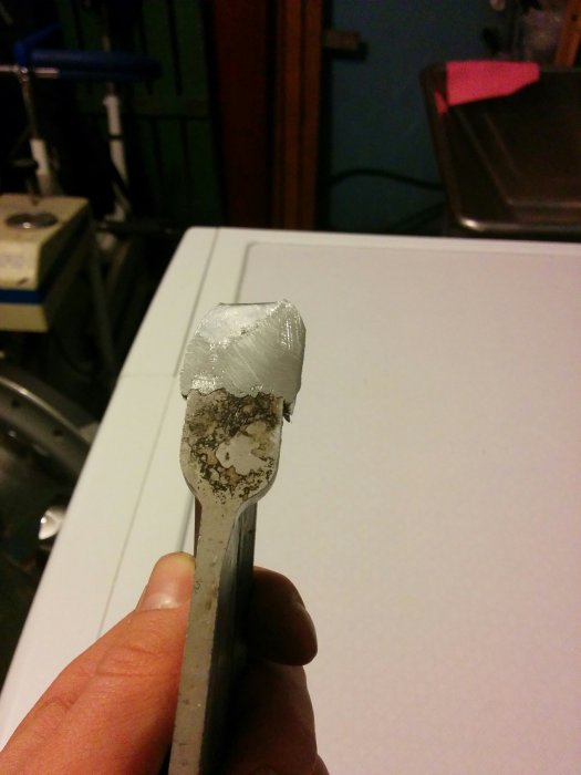 Hand som håller en metallbit med avlångt hål och smält material på toppen efter bearbetning.