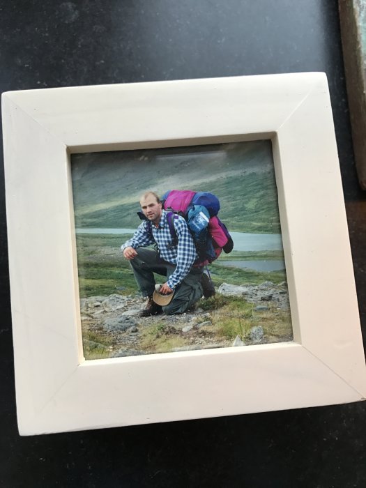 Person på huk i fjällterräng med ryggsäck, fotografi i ram på bord.
