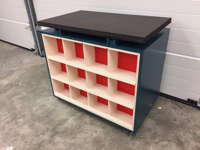 Ett blått arbetsbord med svart topp kombinerat med birgplywood hyllor och röda förvaringsfack.