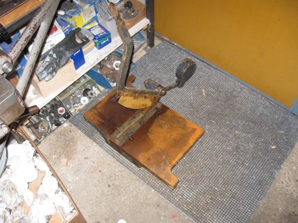 Gammal gjutjärnsklipp på ett verkstads-golv omgeben av verktyg och skruvar.