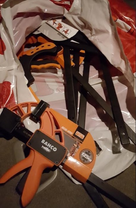 Flera nya tvingar ligger på ett golv med en vit plastpåse, inklusive orange 125KG BAHCO-tvingar och svarta 60 KG-tvingar.