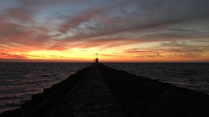 Solnedgång över havet med silhuett av pir och fyr i motljus.