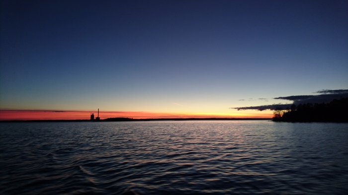 Solnedgång över havet med siluett av en byggnad och träd vid horisonten.
